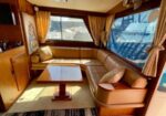 Cozy boat cabin lounge with L-shaped brown leather seating around a wooden table and large windows showing a marina outside.