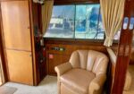 Wood-paneled boat cabin featuring a beige leather armchair and a large window with curtains; marina visible outside.