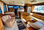 Cozy boat cabin interior with a beige L-shaped sofa around a wooden dining table, a beige armchair, wood-panelled walls, and a galley area with microwave and cupboards; windows with curtains reveal a blue exterior.