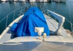 Bow of a boat with blue tarp covering equipment on deck, marina with moored boats in the background.