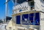 Rear deck of a sailboat with stairs to the upper cockpit, blue panels, and intricate rigging against a bright blue sky.