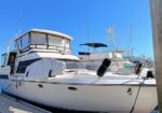 White motor yacht moored at a marina, with a covered upper deck, protective canvas covers, and fenders along the hull under a clear blue sky.