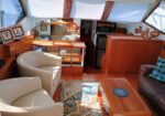 Sunlit yacht interior featuring white curved chairs, wooden cabinetry, a central coffee table, and a patterned rug.