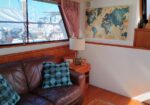 Cozy boat cabin seating area with a brown leather sofa and turquoise patterned cushions, a lamp on a wooden side table, and a world map on the wall; marina visible through the window.