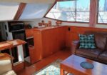 Cozy yacht salon with wooden cabinetry, a brown leather sofa, and a coffee table; harbor seen through large windows.