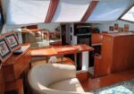 Cozy wooden boat cabin interior with a white curved lounge chair, a desk, framed pictures, a small TV, and skylights with red curtains.