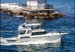 White motorboat cruising through blue water beside a rocky breakwater and a small white building on a jetty with arches