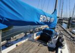 Blue sail cover on a sailboat at a marina, with rigging and masts in the background and calm water nearby.