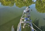Close-up of a metal boat bow and rigging over calm green water with tree reflections in the background, taken from above.