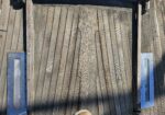Blue rectangular metal mailbox slot mounted on weathered wooden deck planks, with visible screws and a rusted strip nearby.