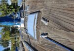 Deck of a sailboat with wooden planks, blue sail cover, and metal fittings; other boats docked in a calm marina in the background.
