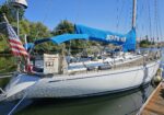 Sailboat docked at a marina with a blue sail cover reading BONES VIII, American flag at the stern, and calm water nearby on a sunny day.
