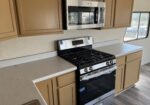 Stainless steel microwave above a gas range in a beige kitchen with speckled countertops and a window nearby.