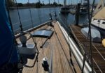 Wooden sailboat deck in a calm marina, with rigging, a hatch, and nearby moored boats visible along the dock