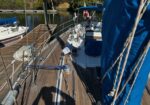 Deck of a sailboat at a marina with ropes, winches, and a blue sail cover; calm water and other boats in the background.