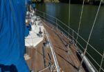 Sailboat deck with wooden planks, winches and ropes, and a blue cover, beside calm water on the right.