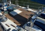 Teak deck of a sailing yacht with winches, ropes, and a hatch, water visible in the background.