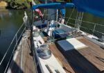 Sailboat deck with wooden planks, winches, lines, and a blue canopy, moored on calm water nearby