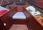 Interior of a small wooden boat with burgundy panels and a narrow central aisle, light blue cushion on a bench, and a white circular hatch cover on the right side.