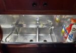 Double-basin stainless steel kitchen sink under a dark wooden cabinet with a chrome faucet in the center and cleaning bottles on the right.