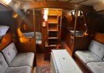 Cozy wooden sailboat cabin with gray upholstered benches, a central ladder, and a small galley on the right; warm lighting.