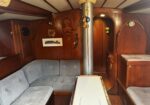 Wood-paneled boat cabin with a gray sectional sofa along the left wall and a central metal support column; nautical decorations on the wall, including a carved tikis and a small mask, and a clock on the right wall.