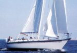 White sailboat with sails raised gliding on blue water, crew on deck amid a sunny sea day?