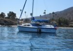 Sailboat named 'Corsair' anchored in calm blue water near a rocky shore with palm trees and hills in the background.