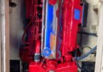 Red engine with blue valve cover in a wooden engine bay, wires and hoses visible around it