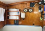 Navigation station in a wood-paneled boat cabin: shelf with manuals, two gauges, a wall lamp, radio, and a cushioned bench below.