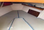 V-shaped boat bunk with light gray cushions and blue piping, under a wooden cockpit lip; storage shelves along the sides.