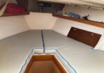 Interior of a sailboat cabin with a V-shaped double berth, light gray cushions with blue piping, and wooden storage panels.