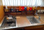 Compact galley with a kettle on a stove, a stainless sink, and a shelf holding colorful cups and jars under a window.