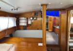 Wood-paneled sailboat cabin with a polished wooden table, bench seating, navigation gauges, and a doorway to the forward cabin; blue hatch overhead and a coiled pink rope nearby.