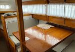 Compact wooden boat cabin with a polished table, built-in bench seating, and a small bed along the left; white curtains over narrow windows.