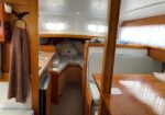 Interior of a small sailboat cabin: wood paneling, U-shaped seating with blue cushions, a wooden table, and a forward berth; small windows let in light.