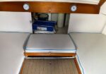 Interior of a small boat cabin showing cushioned U‑shaped seating and a fold‑down berth, with an open hatch revealing a blue cooler and storage area beneath; gauges mounted on a wooden beam above.