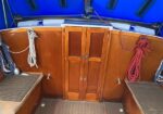 Sailboat cockpit interior with a central wooden cabinet, cushioned benches on both sides, and coiled white and red ropes hanging along the wood panels under a blue canopy.