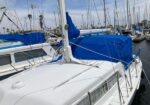 White sailing yacht docked in a marina with blue canvas covers over the cockpit, surrounded by masts and neighboring boats under a clear sky.