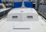 White sailboat deck with two small rectangular porthole-style windows and a blue cover at the stern, moored in a marina with other boats and masts in the background.