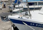 Bow of a white sailboat with a blue stripe tied at a marina, showing anchoring hardware and lines; other boats are visible in the background.