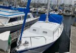 White and blue sailboat moored at a marina, with masts and other boats in the background, and a blue sail cover over the cockpit.