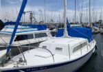 White sailboat docked in a marina with blue canvas covers, surrounded by other moored boats on calm water.