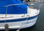 Blue-caded sailboat with a blue cover moored at a marina, white hull with a blue stripe and the name 'Corsair' visible on the bow, tied to dock with fenders nearby.