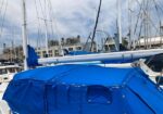 Blue boat cover secured over a sailboat at a marina, with masts and other boats in the background.