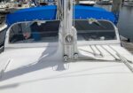 Bow of a white sailboat with blue dodger at a marina, showing rigging, mast base, and winches on the foredeck.