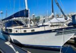 Sailboat with white hull and blue trim docked at a marina, blue canvas cover over the cockpit and masts in the foreground against a clear blue sky.