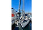 Sailboat docked at a marina with stainless stern rails, rigging, and other boats in the bright blue sky backdrop.