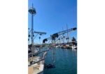 Marina scene with moored sailboats along a calm blue harbor; foreground shows a stainless steel boat davit and railing against a bright sky.