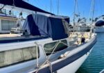 Sailboat docked at a marina with a navy canvas cover over the cockpit, white hull with a dark blue stripe, stainless rails, and mooring lines by calm water with other boats in the background.